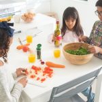 Three children preparing a fresh vegetable salad together in a bright kitchen.