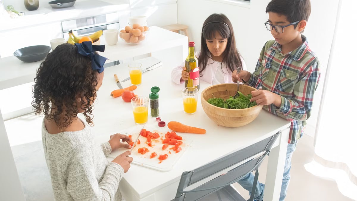Three children preparing a fresh vegetable salad together in a bright kitchen.